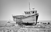A large, weathered fishing boat rests on a pebbled shore with a smaller sailboat in the background near a calm sea. The scene is depicted in black and white, enhancing the rustic and abandoned vibe of the location.