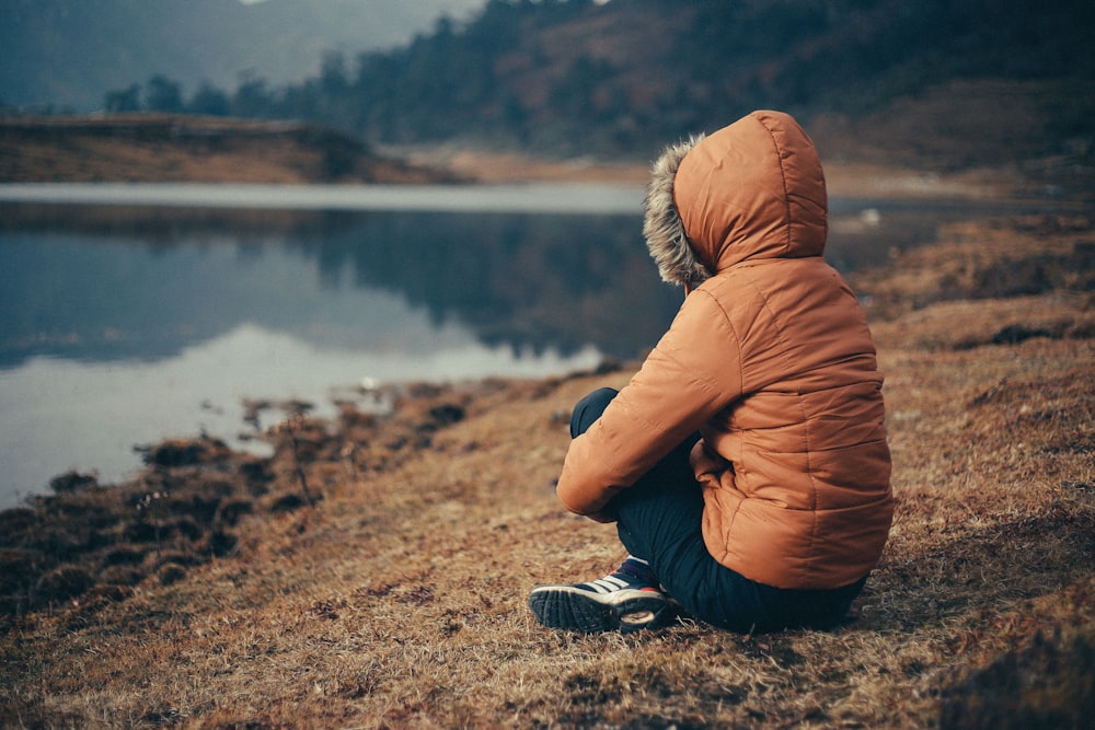 woman in orange parka sits on bank of lake