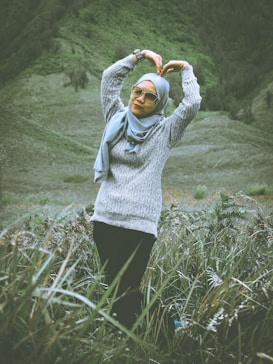 A person wearing sunglasses and a hijab stands in a grassy field with arms raised in a heart shape above their head. They are wearing a gray sweater and black pants, against a backdrop of rolling green hills.