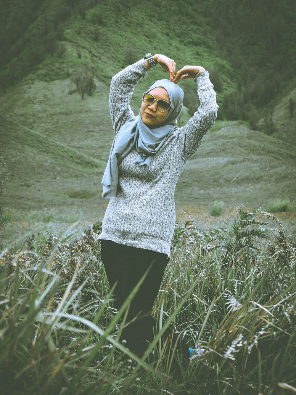 Mujer con suéter de punto gris y pantalones negros haciendo un signo de corazón