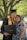 A graduate in a cap and gown is kissing a woman on the forehead as they both pose happily in front of a tree. The graduate is holding a diploma case, indicating a recent graduation ceremony.