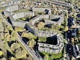 Aerial view of a large residential complex with multiple interconnected buildings arranged in a geometric pattern. The buildings are surrounded by trees and greenery, with roads and additional houses in the vicinity. The overall layout is sprawling and urban, with a mix of concrete and natural elements.