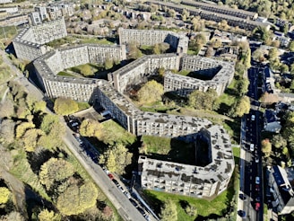 Aerial view of a large residential complex with multiple interconnected buildings arranged in a geometric pattern. The buildings are surrounded by trees and greenery, with roads and additional houses in the vicinity. The overall layout is sprawling and urban, with a mix of concrete and natural elements.