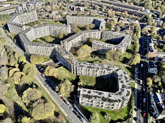Aerial view of a large residential complex with multiple interconnected buildings arranged in a geometric pattern. The buildings are surrounded by trees and greenery, with roads and additional houses in the vicinity. The overall layout is sprawling and urban, with a mix of concrete and natural elements.
