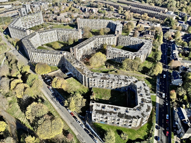 Aerial view of a large residential complex with multiple interconnected buildings arranged in a geometric pattern. The buildings are surrounded by trees and greenery, with roads and additional houses in the vicinity. The overall layout is sprawling and urban, with a mix of concrete and natural elements.