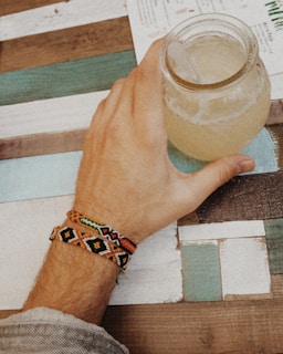 A hand holding a glass jar filled with a light brown liquid sitting on a striped wooden table. The hand features colorful woven friendship bracelets. The background includes a menu or leaflet partially visible on the table.