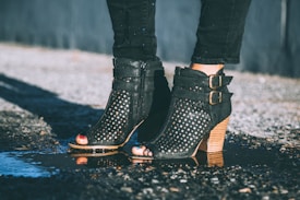 A pair of stylish black ankle boots with cut-out designs and buckles is being worn with black jeans. The boots are shown standing on a wet surface, possibly a street, with light reflecting off the puddle beneath them.
