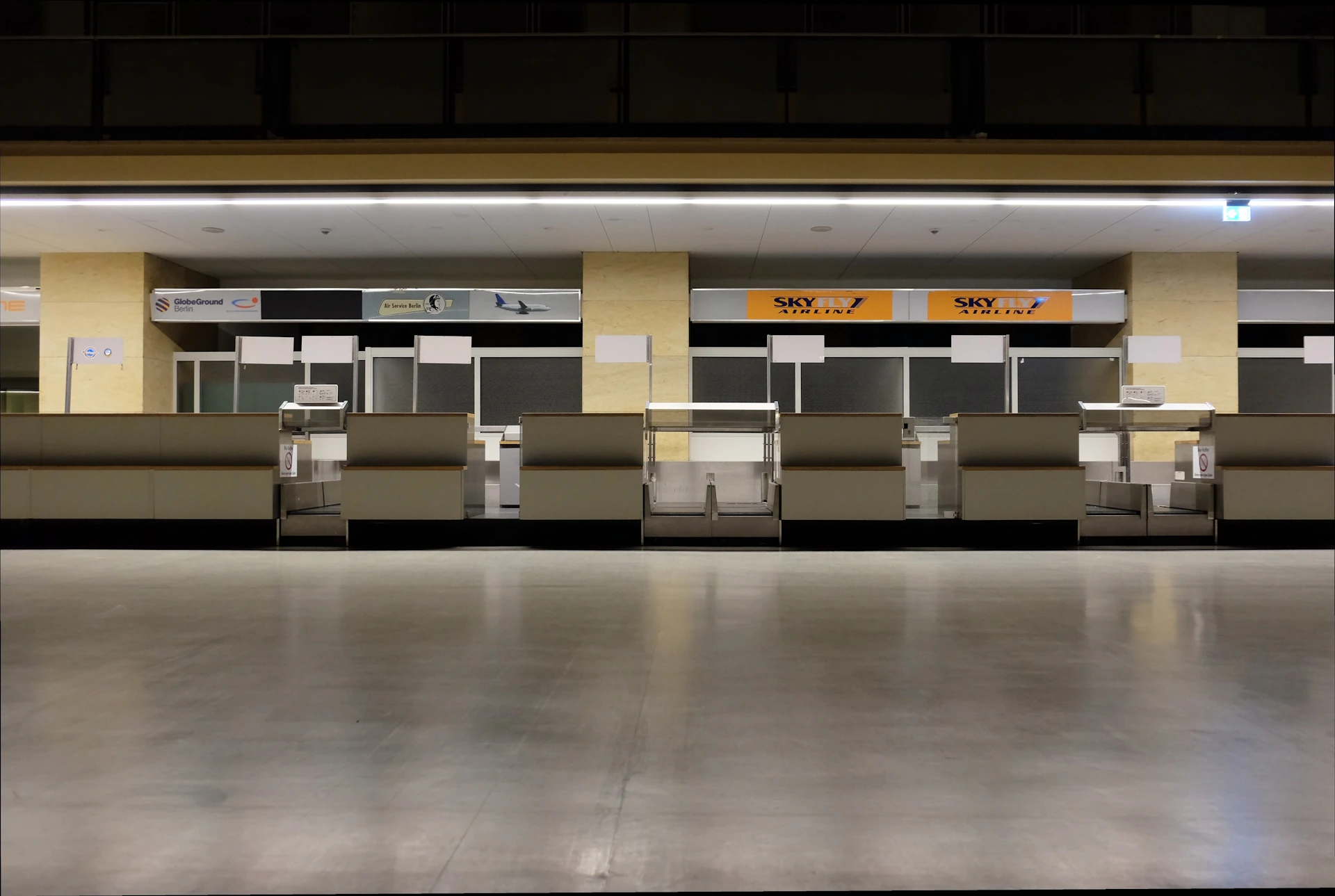 A row of empty airport check-in counters with closed blinds. The counters have signs for different airlines, and the lighting is subdued. The floor is polished and reflective, giving the area a clean and modern feel.