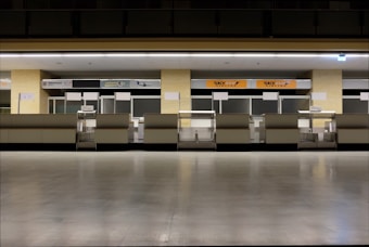 A row of empty airport check-in counters with closed blinds. The counters have signs for different airlines, and the lighting is subdued. The floor is polished and reflective, giving the area a clean and modern feel.