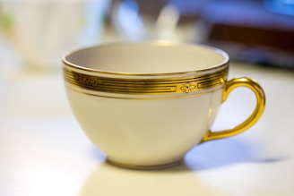 A close-up image of an elegant white teacup with intricate floral and line designs in gold along the rim and on the handle, which is also gold-colored. The teacup is placed on a light-colored surface, and the background appears softly blurred, suggesting a depth of field effect.