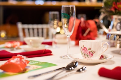 A beautifully arranged table set for a formal dining event, featuring a floral teacup on a saucer, a champagne flute, and elegantly folded red napkins. The table is decorated with silverware, a table ornament, and a blurred background indicating a festive or elegant setting.