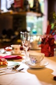 A beautifully arranged dining table setting featuring an ornate teacup and saucer with floral patterns, a champagne flute, and assorted cutlery. The setting includes vibrant touches like red napkins and colorful gift wrapping, contributing to an elegant atmosphere.