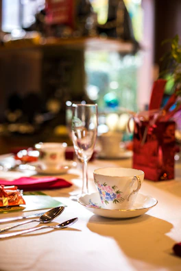 A beautifully arranged dining table setting featuring an ornate teacup and saucer with floral patterns, a champagne flute, and assorted cutlery. The setting includes vibrant touches like red napkins and colorful gift wrapping, contributing to an elegant atmosphere.
