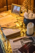 A table setting featuring a variety of items arranged on a shiny gold surface. There are open books, a newspaper with the word 'SHRIEKING SHACK,' a white candle on a black holder, and a wand placed diagonally across the newspaper. A framed photo and a small decorative tree enhance the setup, along with a black cauldron filled with artificial snow.
