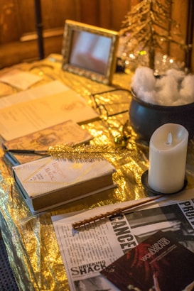 A table setting featuring a variety of items arranged on a shiny gold surface. There are open books, a newspaper with the word 'SHRIEKING SHACK,' a white candle on a black holder, and a wand placed diagonally across the newspaper. A framed photo and a small decorative tree enhance the setup, along with a black cauldron filled with artificial snow.