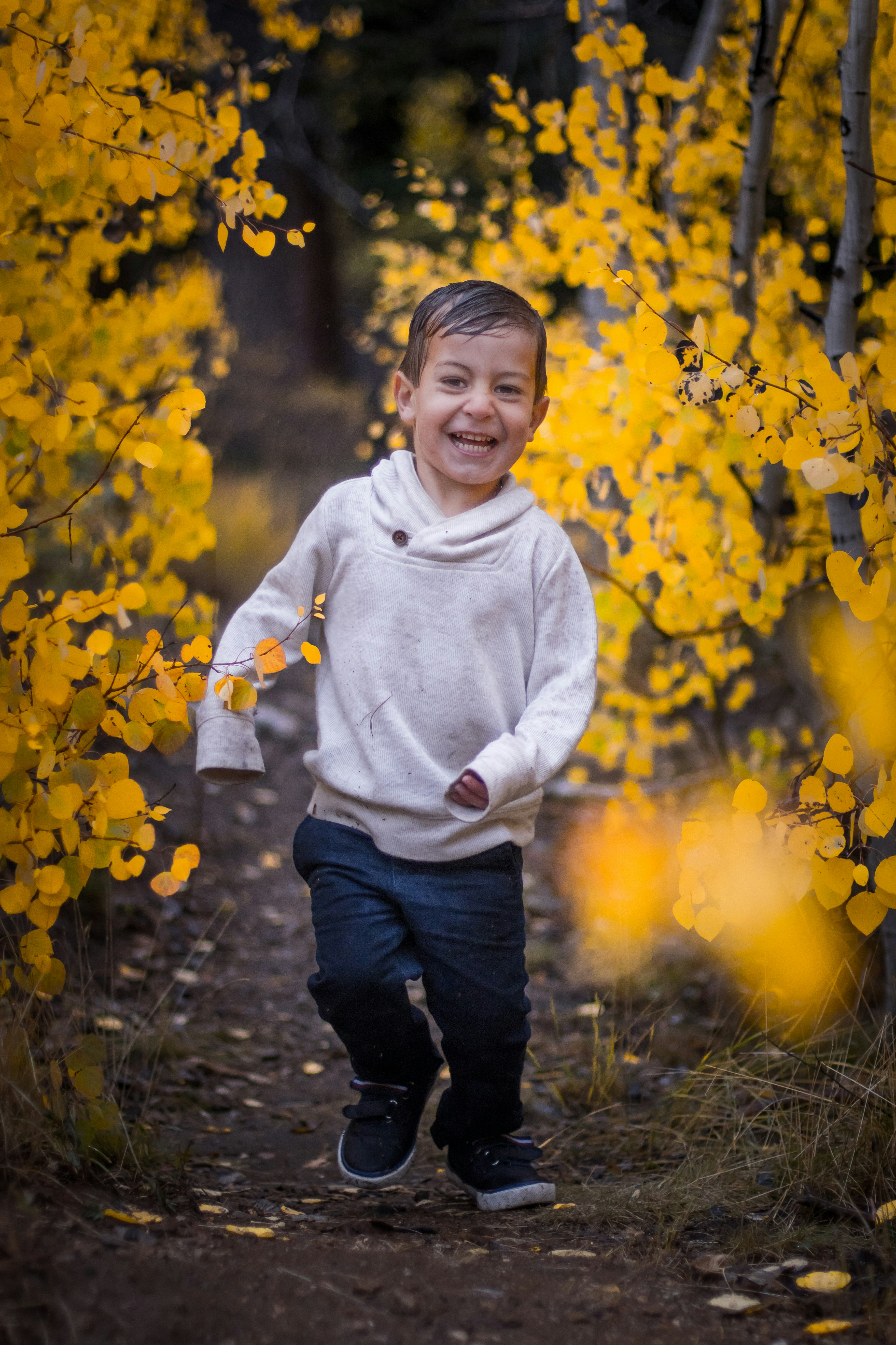 Junge in weißem Pullover-Kapuzenpullover und blauer Jeans läuft auf unbefestigtem Weg neben gelben Blättern