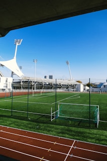 A green soccer field surrounded by a running track under bright floodlights, with a clear blue sky in the background. There are goalposts and a fenced area, with a modern building and tall towers visible at a distance.