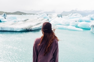 A person with long hair wearing a dark knit cap and a jacket is standing near a body of water with large icebergs floating nearby. The water has a serene, turquoise hue and is surrounded by distant, misty mountains in the background. The view is calm and expansive, emphasizing the natural beauty of the icy landscape.