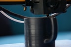 A 3D printer nozzle is actively printing a black cylindrical object with a handle, possibly a cup. The scene shows a close-up of the printing process, highlighting the detail of the extruding filament.