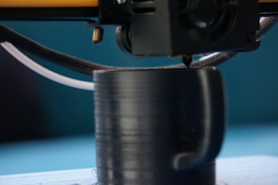 A 3D printer nozzle is actively printing a black cylindrical object with a handle, possibly a cup. The scene shows a close-up of the printing process, highlighting the detail of the extruding filament.