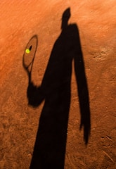 A shadow of a person holding a tennis racket is cast on an orange-brown clay tennis court. A tennis ball is visible in mid-air near the racket.