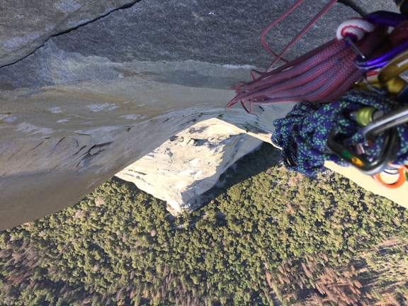 A climber is suspended on a vertical rock face, with various climbing gear such as ropes and carabiners visible in the foreground. Below is a vast view of a forested landscape, providing a dramatic perspective of height and exposure.
