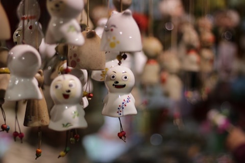A collection of small porcelain or ceramic wind chimes are hanging closely together. Each chime has a unique face and design, featuring painted expressions and colorful patterns. The chimes are suspended with strings and have small beads tied near the bottom. The background is blurred, with a bokeh effect highlighting the chimes in the foreground.