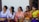 A group of women are sitting together, dressed in traditional colorful attire. They are holding small woven baskets and seem to be engaged in a conversation or an activity involving a phone. The background includes a wooden wall and a white section, suggesting an outdoor setting.