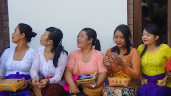A group of women are sitting together, dressed in traditional colorful attire. They are holding small woven baskets and seem to be engaged in a conversation or an activity involving a phone. The background includes a wooden wall and a white section, suggesting an outdoor setting.