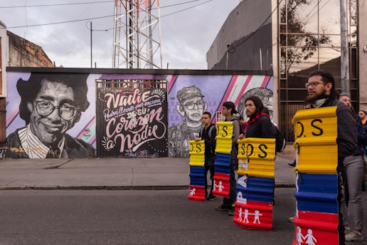 A colorful mural on a city street features a large portrait of a man with glasses and text that reads 'Nadie podrá llevar por encima de su corazón a nadie.' Several people stand in front of the mural, each holding shields painted with red, yellow, and blue colors and the letters 'S.O.S'. The setting is urban with surrounding modern buildings and a cloudy sky.