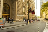 A busy urban street features a grand building with the address 26 Broadway. People are walking near the entrance. On the sidewalk, there are several street vendors selling various items, including food from a cart. Banners for the New York Film Academy School of Film and Acting hang from a lamppost. A white van is parked on the street, and trees with green foliage can be seen in the background.