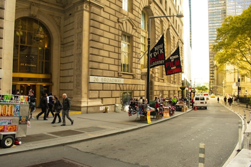 A busy urban street features a grand building with the address 26 Broadway. People are walking near the entrance. On the sidewalk, there are several street vendors selling various items, including food from a cart. Banners for the New York Film Academy School of Film and Acting hang from a lamppost. A white van is parked on the street, and trees with green foliage can be seen in the background.