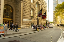A busy urban street features a grand building with the address 26 Broadway. People are walking near the entrance. On the sidewalk, there are several street vendors selling various items, including food from a cart. Banners for the New York Film Academy School of Film and Acting hang from a lamppost. A white van is parked on the street, and trees with green foliage can be seen in the background.
