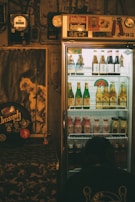 A dimly lit room features a refrigerated display case filled with assorted drinks, including bottles of beer and other beverages. The fridge is topped with various stickers and decor. To the left, a poster and some musical equipment can be seen, adding a retro vibe to the ambiance. A person is crouched near the bottom of the fridge, perhaps examining the drinks.