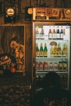 A dimly lit room features a refrigerated display case filled with assorted drinks, including bottles of beer and other beverages. The fridge is topped with various stickers and decor. To the left, a poster and some musical equipment can be seen, adding a retro vibe to the ambiance. A person is crouched near the bottom of the fridge, perhaps examining the drinks.