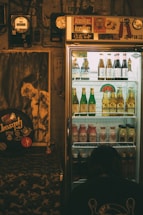 A dimly lit room features a refrigerated display case filled with assorted drinks, including bottles of beer and other beverages. The fridge is topped with various stickers and decor. To the left, a poster and some musical equipment can be seen, adding a retro vibe to the ambiance. A person is crouched near the bottom of the fridge, perhaps examining the drinks.