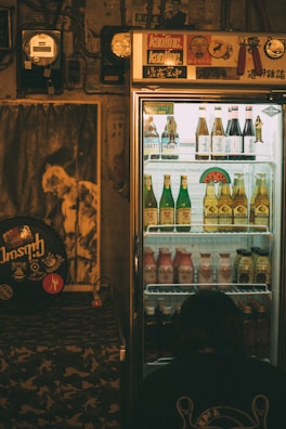 A dimly lit room features a refrigerated display case filled with assorted drinks, including bottles of beer and other beverages. The fridge is topped with various stickers and decor. To the left, a poster and some musical equipment can be seen, adding a retro vibe to the ambiance. A person is crouched near the bottom of the fridge, perhaps examining the drinks.
