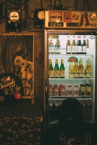 A dimly lit room features a refrigerated display case filled with assorted drinks, including bottles of beer and other beverages. The fridge is topped with various stickers and decor. To the left, a poster and some musical equipment can be seen, adding a retro vibe to the ambiance. A person is crouched near the bottom of the fridge, perhaps examining the drinks.
