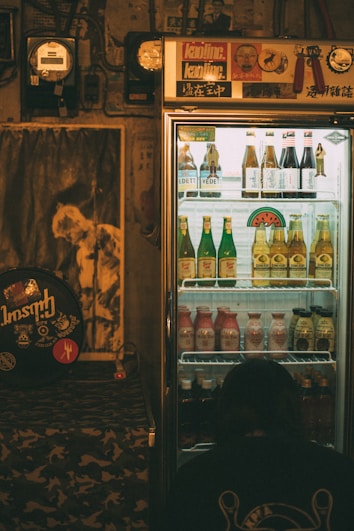 A dimly lit room features a refrigerated display case filled with assorted drinks, including bottles of beer and other beverages. The fridge is topped with various stickers and decor. To the left, a poster and some musical equipment can be seen, adding a retro vibe to the ambiance. A person is crouched near the bottom of the fridge, perhaps examining the drinks.