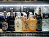 A selection of various bottled beverages are arranged on a refrigerated shelf. The lineup includes multiple types of Asian beers and sake, along with bottles of water. Each bottle displays unique and colorful labels featuring distinct branding and design elements.