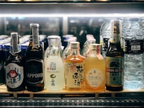 A selection of various bottled beverages are arranged on a refrigerated shelf. The lineup includes multiple types of Asian beers and sake, along with bottles of water. Each bottle displays unique and colorful labels featuring distinct branding and design elements.