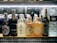 A selection of various bottled beverages are arranged on a refrigerated shelf. The lineup includes multiple types of Asian beers and sake, along with bottles of water. Each bottle displays unique and colorful labels featuring distinct branding and design elements.