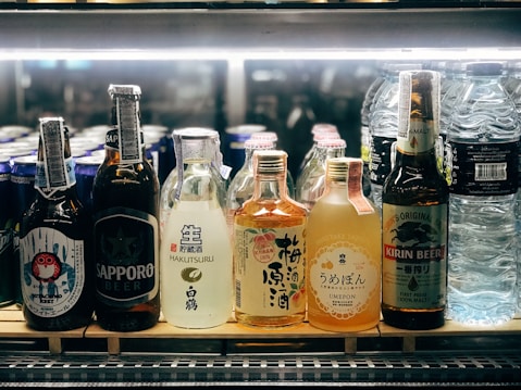 A selection of various bottled beverages are arranged on a refrigerated shelf. The lineup includes multiple types of Asian beers and sake, along with bottles of water. Each bottle displays unique and colorful labels featuring distinct branding and design elements.