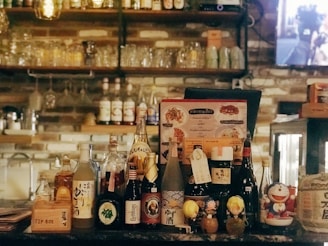 A bar counter filled with various bottles of alcohol including sake and sprite, surrounded by small figurines and a tip box. Behind the counter are shelves with more glasses and bottles, creating a cozy and inviting atmosphere.