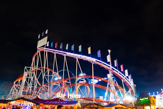A colorful amusement park roller coaster is shown at night, adorned with vibrant lights and flags along its tracks. The roller coaster has loops and steep climbs, with the words 'Bar H&uuml;tte' illuminated on a nearby structure. The scene is bustling with activity and other visible attractions under the dark night sky.