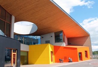 A modern building with a unique architectural design featuring vibrant colors. The structure has a large wooden overhang with an open circular section and glass panels on the upper level. The exterior walls are painted in bright yellow and orange. In front of the building, there are two strollers parked on the paved walkway.