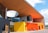A modern building with a unique architectural design featuring vibrant colors. The structure has a large wooden overhang with an open circular section and glass panels on the upper level. The exterior walls are painted in bright yellow and orange. In front of the building, there are two strollers parked on the paved walkway.