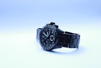 A stylish black wristwatch with a metal strap is positioned against a light background. The watch face displays multiple dials and markings, contributing to a sophisticated design. The lighting adds a cool tone to the watch and its surroundings.