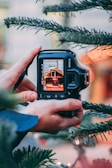 A close-up of a digital camera being held between two hands, capturing an image of a blurred scene that includes soft lights. The frame is surrounded by pine tree branches, suggesting a festive atmosphere. The focus is on the camera's display screen, which clearly shows the scene being photographed.