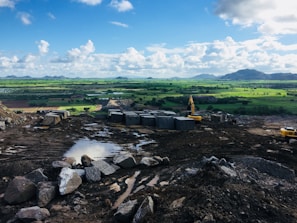 A vast landscape with a construction site in the foreground, featuring piles of rocks and blocks of stone. Construction machinery, including excavators, is visible on the rugged terrain. The background reveals an expansive green field under a sky dotted with fluffy white clouds. Rolling hills are seen in the distance.